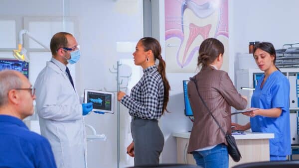 Doctor holding tablet with x-ray showing it to patient while nurse helping woman filling in dental document. Stomatologist presenting dental radiography using modern gadget in stomatological clinic