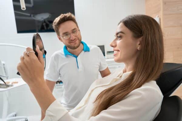 Smiling dental clinic customer looking in hand mirror after procedure in presence of joyous dentist