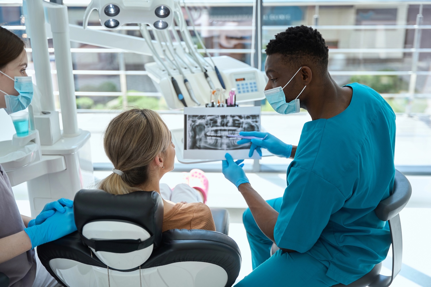 Young doctor in protective mask and blue suit sitting on chair near woman and showing results of treatment