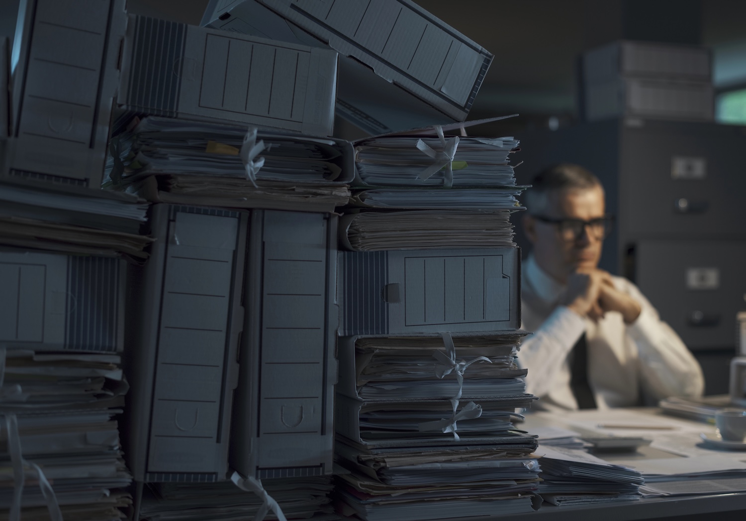 Sad pensive businessman sitting at desk at night and thinking, he is overloaded with paperwork