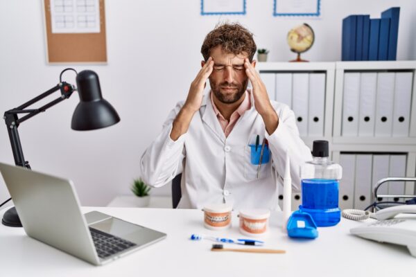 Young hispanic dentist man working at medical clinic with hand on head for pain in head because stress. suffering migraine.