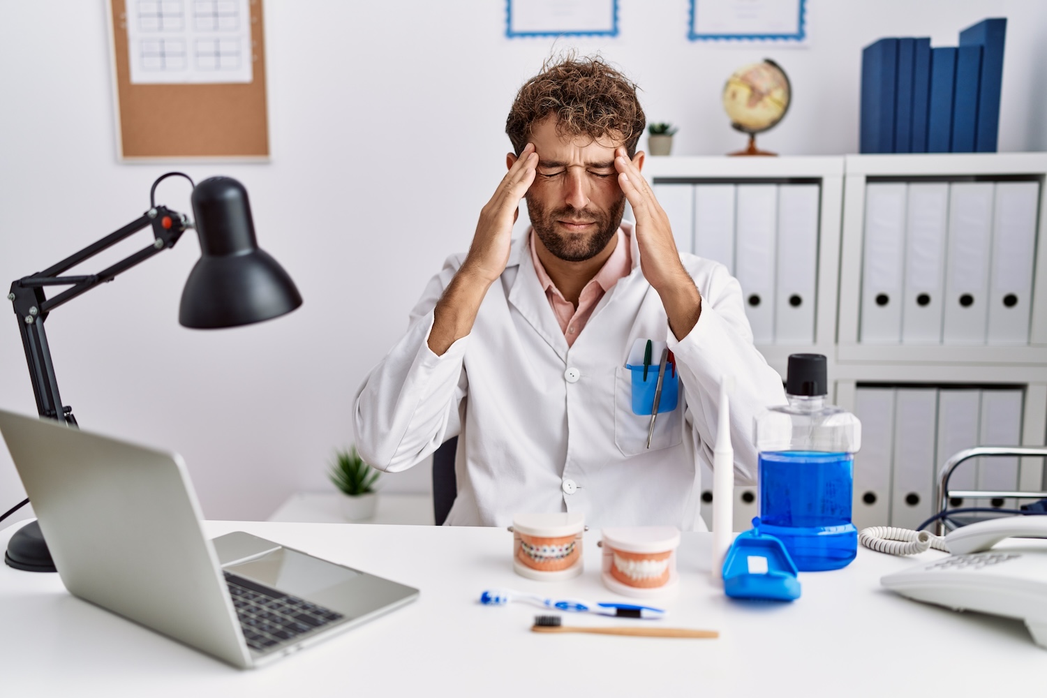 Young hispanic dentist man working at medical clinic with hand on head for pain in head because stress. suffering migraine.