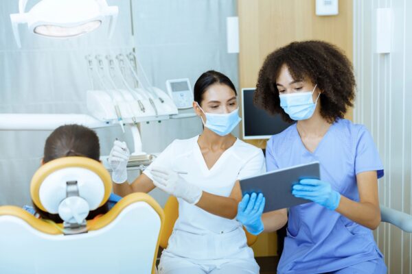 Modern technology and X-rays in dental office. Young european and african american female doctors in protective masks and rubber gloves, treat child in chair and look at tablet in clinic with tools