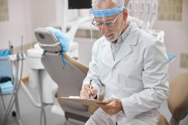 Dentist wearing a full-face shield looking down on his notes while sitting in his clinic
