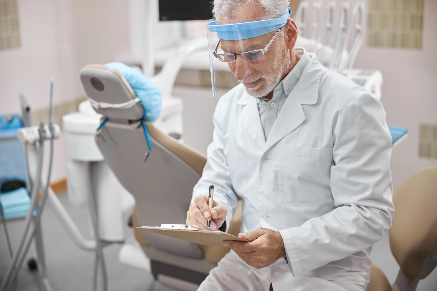 Dentist wearing a full-face shield looking down on his notes while sitting in his clinic