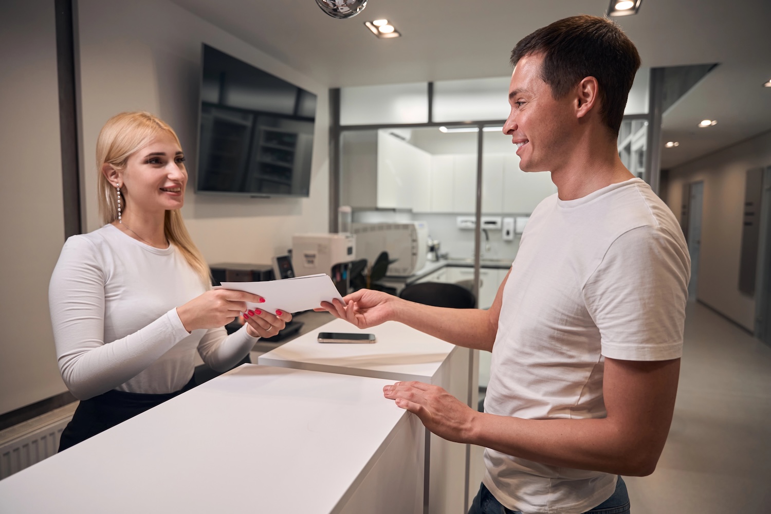 Smiling blond woman worker of dental clinic reception giving the medical records to male client who visiting dental office for routine teeth health check-up